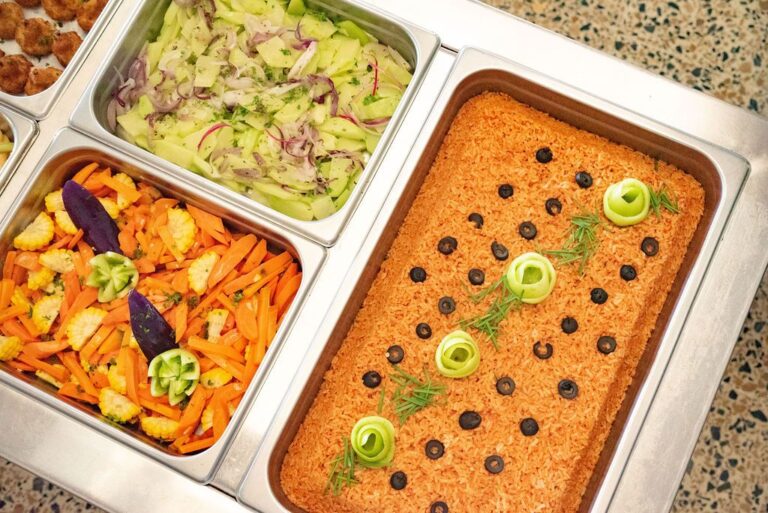 A large tray of seasoned rice with black olives and green onion garnishes is surrounded by two smaller trays containing fresh sliced cucumbers, red onions, and colorful mixed vegetables on Roatan, Honduras.