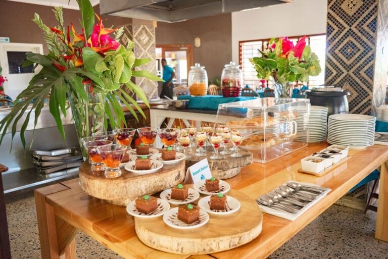 A buffet table on Roatan, Honduras, features colorful floral arrangements, small desserts in glass cups, and assorted pastries arranged on wooden slabs inside a restaurant setting.