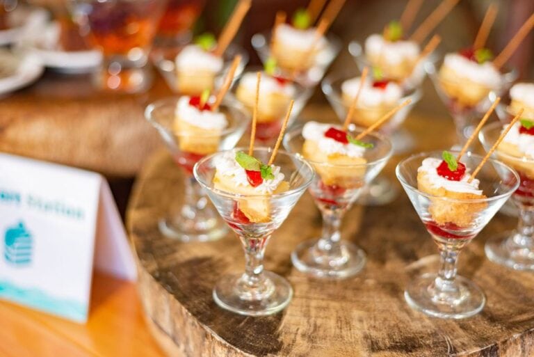Several elegant glass cups filled with layered desserts featuring sponge cake, whipped cream, and red fruit sauce, garnished with mint leaves and a small pastry stick, displayed on a wooden serving board in Roatan, Honduras.