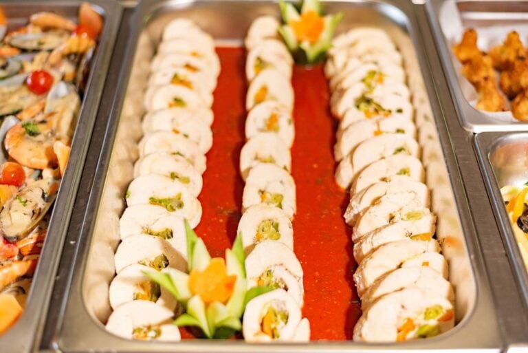 Roatan restaurant buffet in Honduras featuring rows of rolled chicken or fish with green olives and vegetables, garnished with decorative tulip-shaped vegetable flowers, set on a red sauce in stainless steel serving trays.