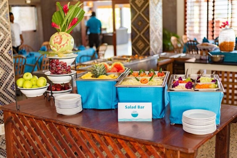 A vibrant salad bar setup with fresh fruits and vegetables on a wooden table inside a restaurant on Roatan, Honduras, featuring options like grapes, apples, pineapple, and colorful salads with tropical decor in the background.