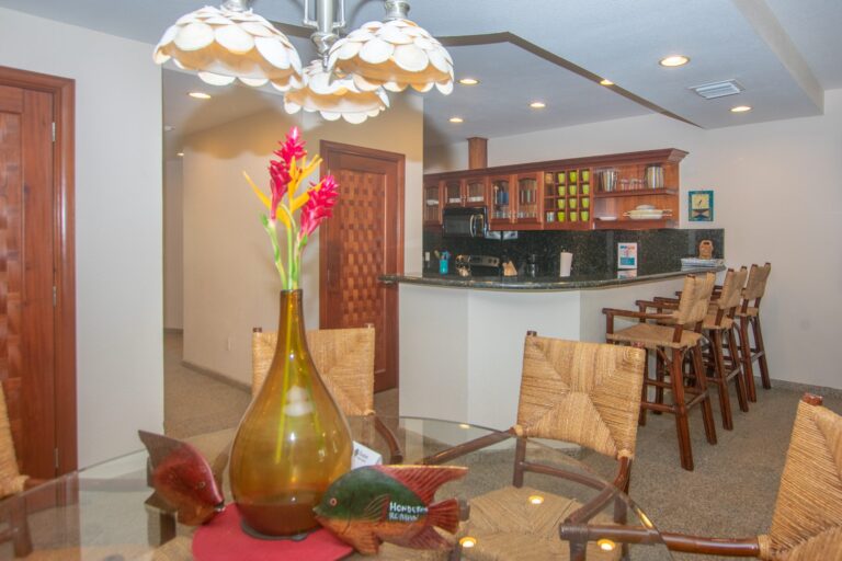 Interior view of a cozy dining area in Roatan, Honduras, featuring a wooden kitchen with open shelves displaying bright green cups and glassware, a bar counter with woven chairs, and decorative elements including a shell pendant light and vibrant tropical flowers.