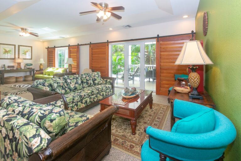 Living room in a tropical-style house on Roatan, Honduras, featuring floral-patterned sofas, wooden accents, and large glass doors opening to a lush outdoor balcony with jungle view.