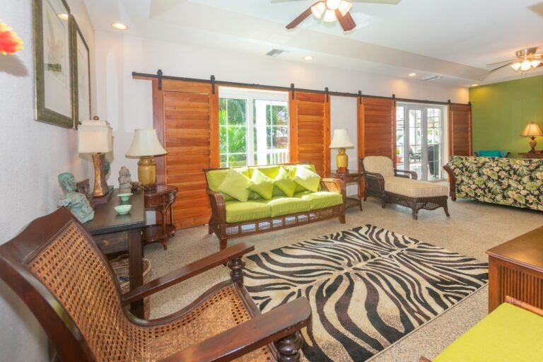 Living room in a Caribbean-style home on Roatan, Bay Islands, Honduras, with wooden sliding barn doors, tropical-themed furniture, and large windows overlooking lush greenery.