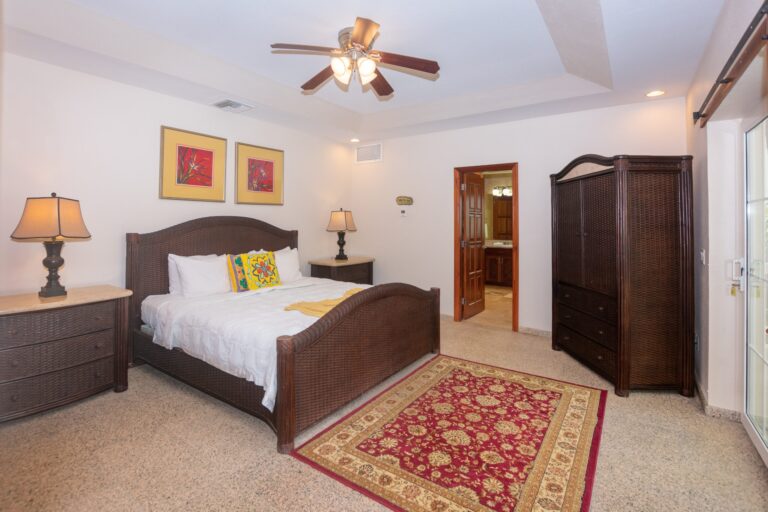 A cozy bedroom in a Roatan, Honduras hotel featuring a wicker bed with white bedding and a colorful decorative pillow, flanked by matching bedside tables and lamps, with artwork on the wall and a wooden armoire on the right side of the room.
