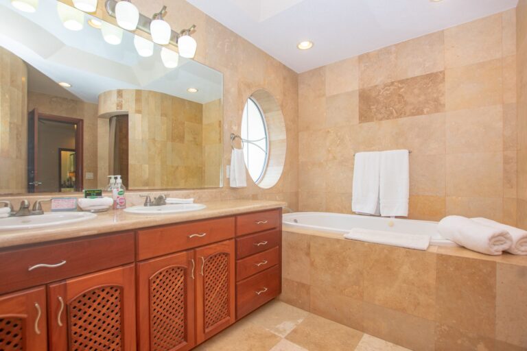 A spacious bathroom on Roatan with beige tiled walls, a large mirror, dual sinks, and a round window with an ocean view. The bathroom features a wooden vanity with ample storage, a white bathtub, and modern lighting fixtures, reflecting the upscale accommodations common on Honduras's Roatan island.