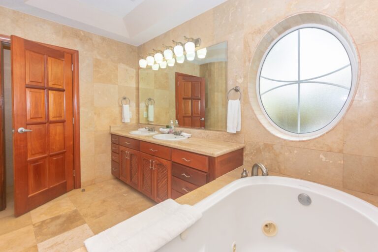 A spacious bathroom in Roatan, Honduras featuring a double sink vanity with a large mirror and modern light fixtures, a round frosted window allowing natural light, and a marble countertop with toiletries, all surrounded by beige tiled walls and floors.