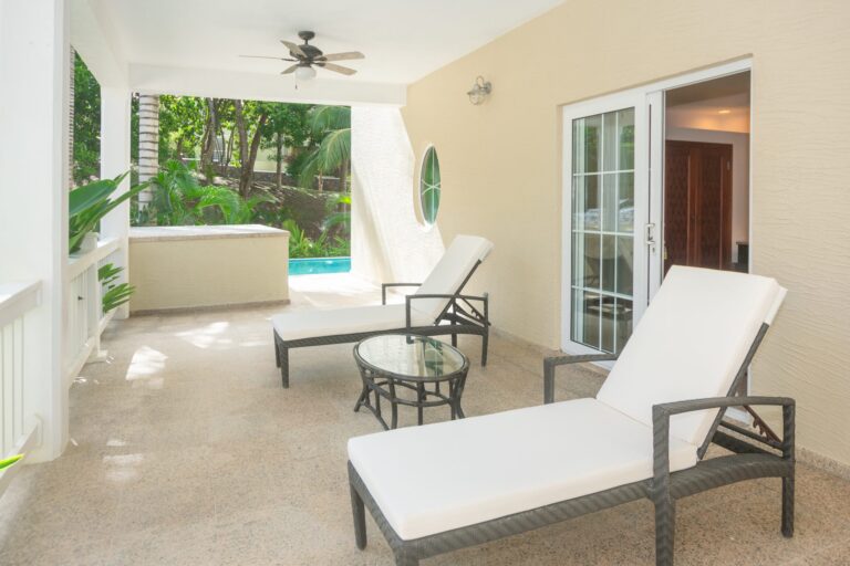 Covered outdoor patio in a Honduran house with white lounge chairs, a glass-topped table, ceiling fan, and a view of lush tropical trees surrounding the property on Roatan in the Bay Islands Honduras.