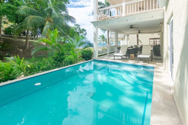 A luxurious swimming pool surrounded by tropical palm trees and a shaded outdoor seating area at a beachfront property on Roatan in Honduras with ocean views in the background.