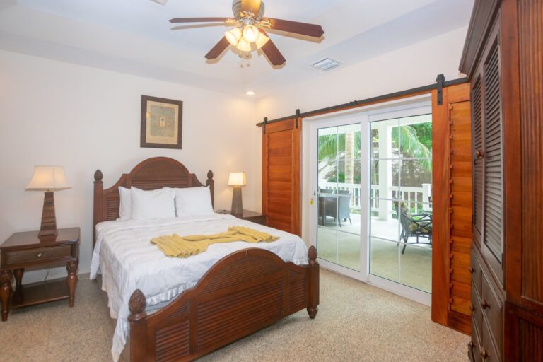 A cozy bedroom with a wooden bed frame, white bedding, and a yellow blanket, featuring sliding glass doors that open to a balcony with outdoor seating and lush green palm trees, typical of Roatan's tropical atmosphere in Honduras.