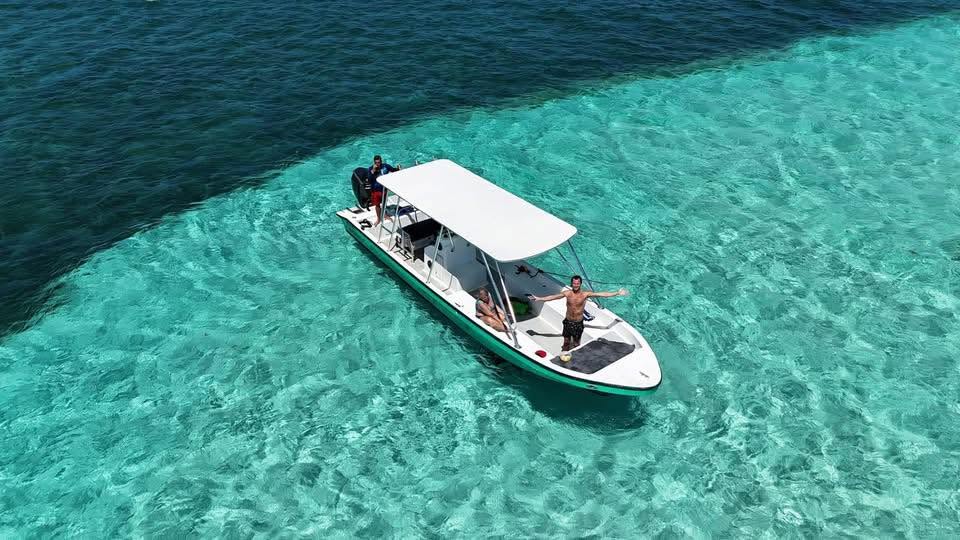 Three people enjoying a boat cruise in the clear turquoise waters near Roatan, Honduras, with a lush island shoreline visible in the distance.