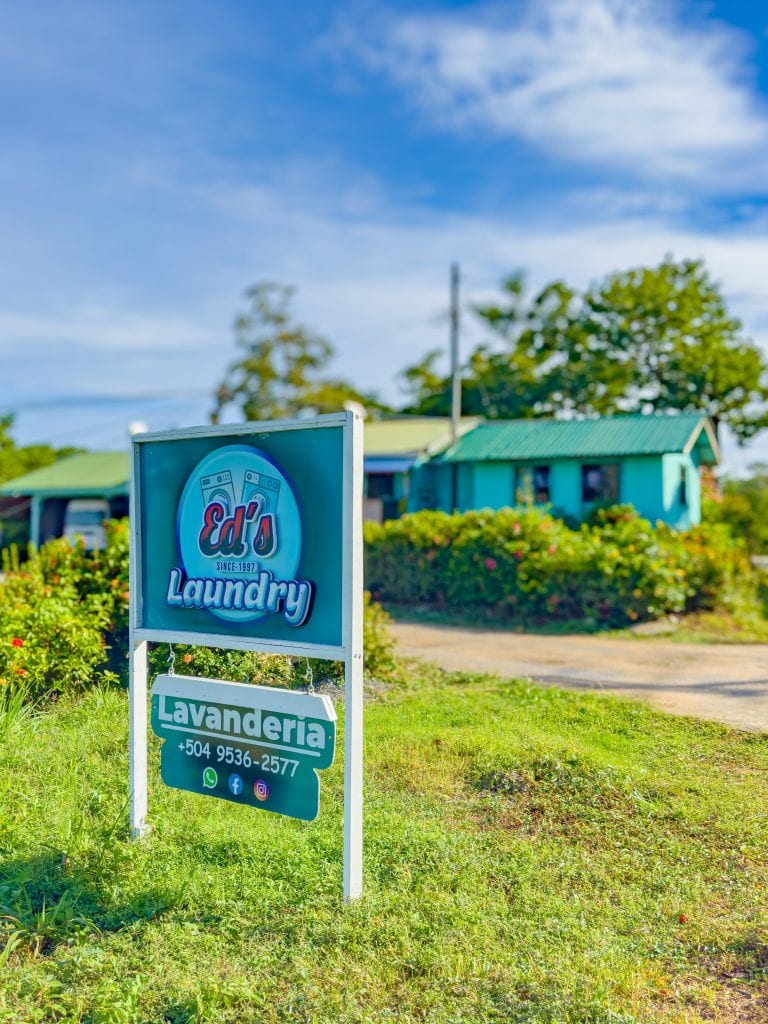 A blue and white sign for Ed's Laundry, showing contact information and social media icons, is situated on a grassy area with a tropical neighborhood of colorful buildings, trees, and a clear blue sky in Roatan, Honduras.
