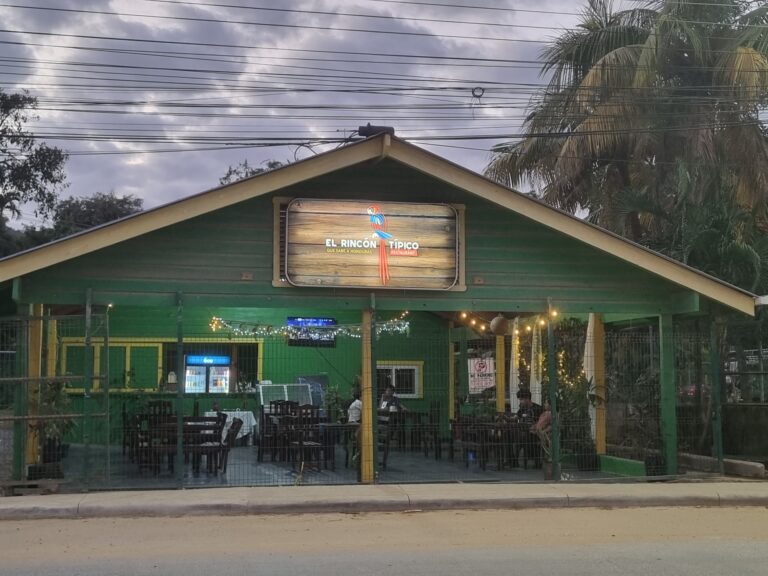 El Rincón Típico is a Honduras restaurant in Roatan with an outdoor seating area enclosed by a green fence, featuring tables with white tablecloths and string lights, set against a cloudy sky with palm trees and electrical wires overhead.
