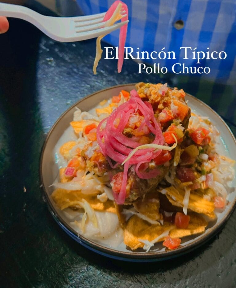 A plate of traditional Honduran pollo chuco with shredded cheese, pickled red onions, and fresh vegetables is shown on a black table, with a fork lifting some of the dish in a Roatan restaurant.