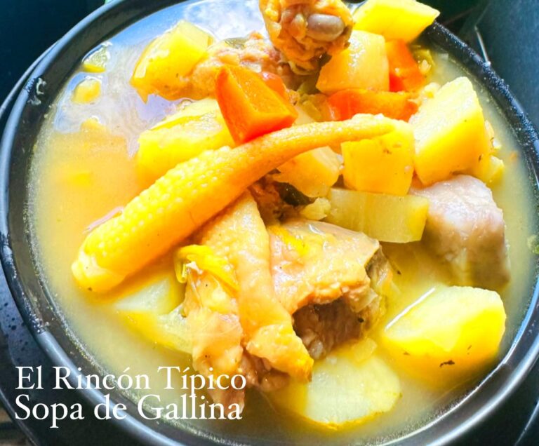 A bowl of El Rincón Típico Sopa de Gallina featuring chunks of chicken, potatoes, carrots, corn on the cob, and other vegetables in a yellow broth, served on a dark table in Roatan, Honduras.