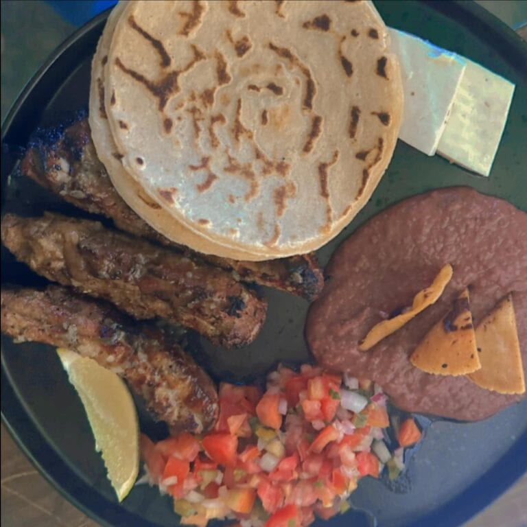 A traditional Honduran meal featuring grilled chicken, spicy sausage, fresh tomato and onion salsa, plantains, a lemon wedge, and a Honduran-style corn tortilla served on a black plate in Roatan, Honduras.