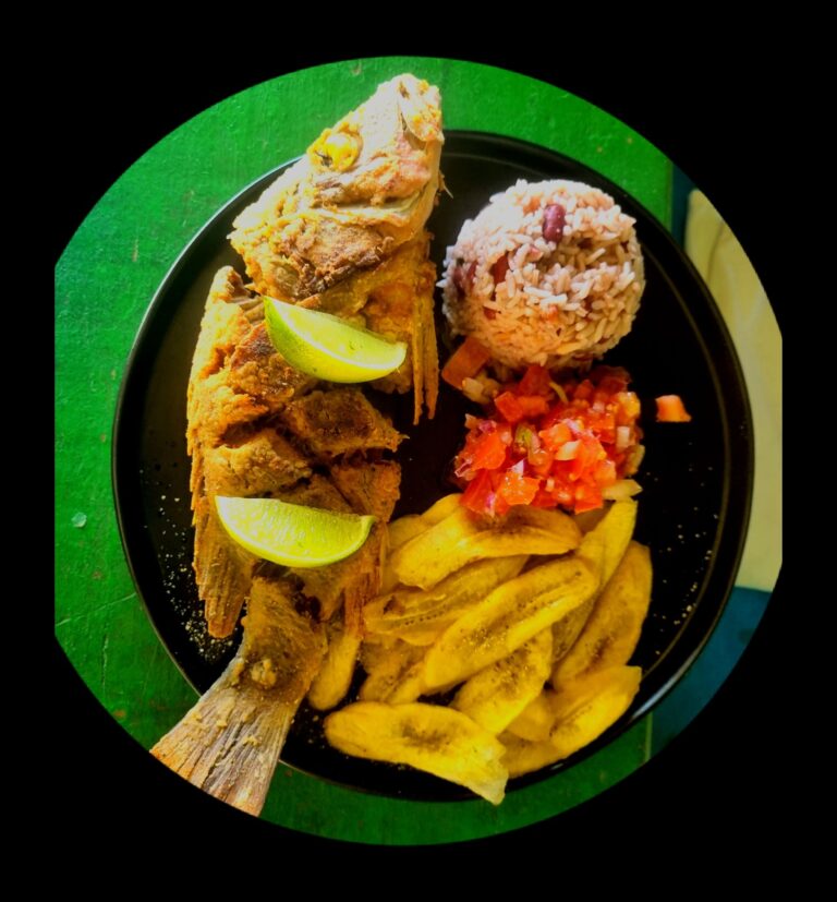 A black plate on a green surface features fried fish garnished with lemon wedges, a mound of rice with red beans, and a serving of vegetable salad with diced carrots and cabbage, representing typical Honduran cuisine on Roatan, Honduras.
