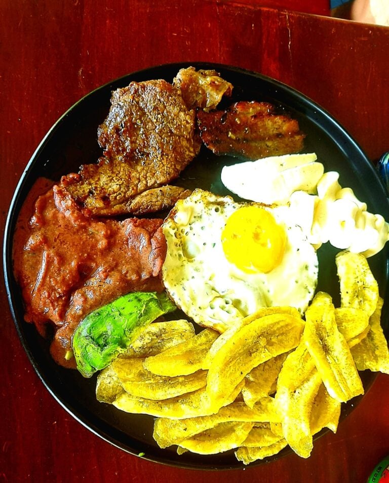 A colorful Honduran plate on Roatan features fried plantains, a sunny-side-up egg, cooked meats, a green pepper, and a small portion of potato salad, all arranged on a black plate set on a wooden table.
