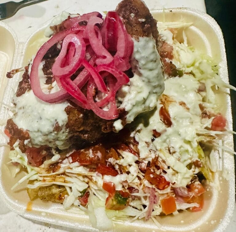 A takeout container displays a traditional Honduran dish with shredded cabbage, diced tomatoes, and sliced onions, topped with a fried meat patty covered in white sauce and garnished with pickled red onions, representing Roatan's cuisine in Honduras.