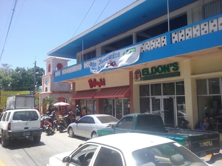A busy street scene in Roatan, Honduras, features parked cars and motorcycles in front of a building with a sign for Eldon's Supermarkets and a banner advertising a seafood restaurant called "Sin Zancudos.