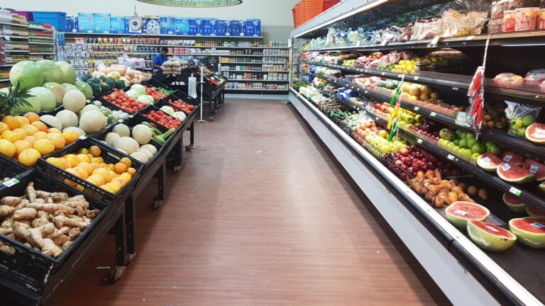 A grocery store aisle in Roatan, Honduras, displays fresh fruits and vegetables on black display bins and shelves, including watermelons, melons, tomatoes, ginger, and apples, with a worker in the background organizing products.