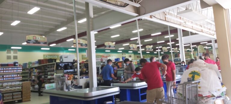 Shoppers at a supermarket checkout counter in Roatan, Honduras, with employees and customers wearing masks, surrounded by grocery shelves and checkout stations inside a bright, modern store.