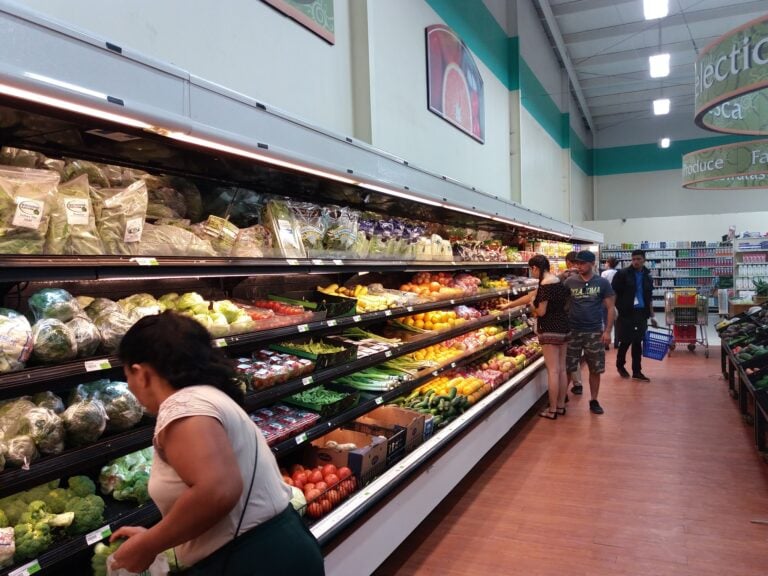 Shoppers browse fresh produce, including lettuce, tomatoes, and zucchinis, on grocery store shelves inside a supermarket on Roatan, Honduras.