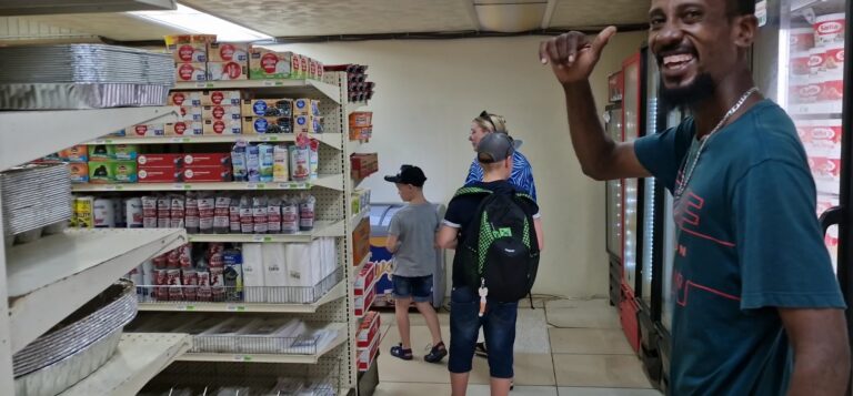 A man smiling and pointing at a grocery store shelf stocked with food items inside a convenience store on Roatan, Honduras, with children shopping and a woman in the background.
