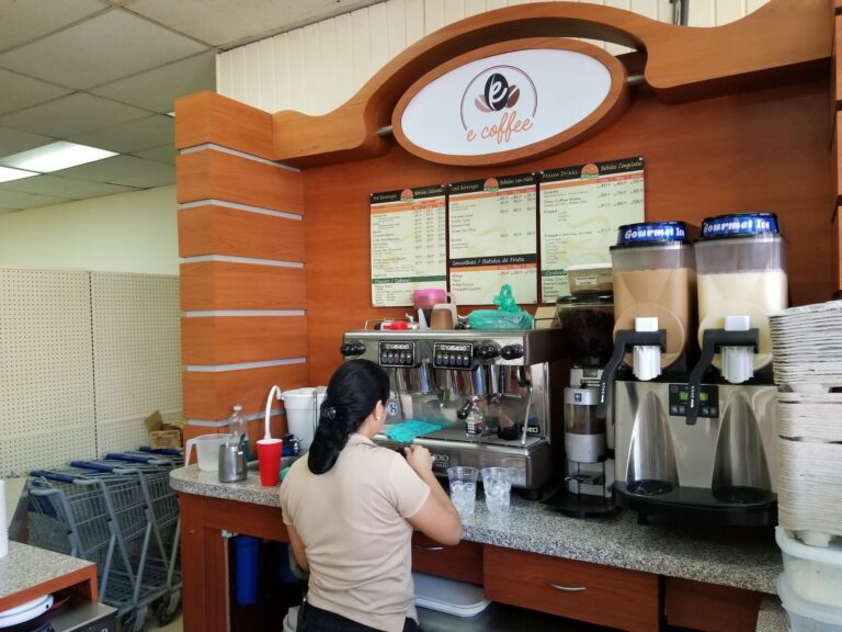 A woman preparing drinks behind the counter at a coffee shop in Roatan, Honduras, with a menu displaying various coffee options and frozen beverages mounted on a wooden wall.