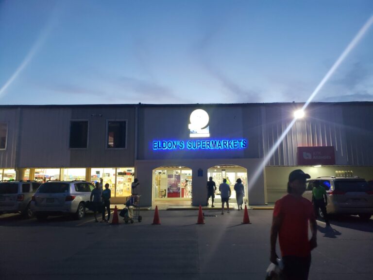 Eldon’s Supermarkets at night in Roatan, Honduras, features a building with a bright neon blue sign and people shopping and walking in the parking lot.