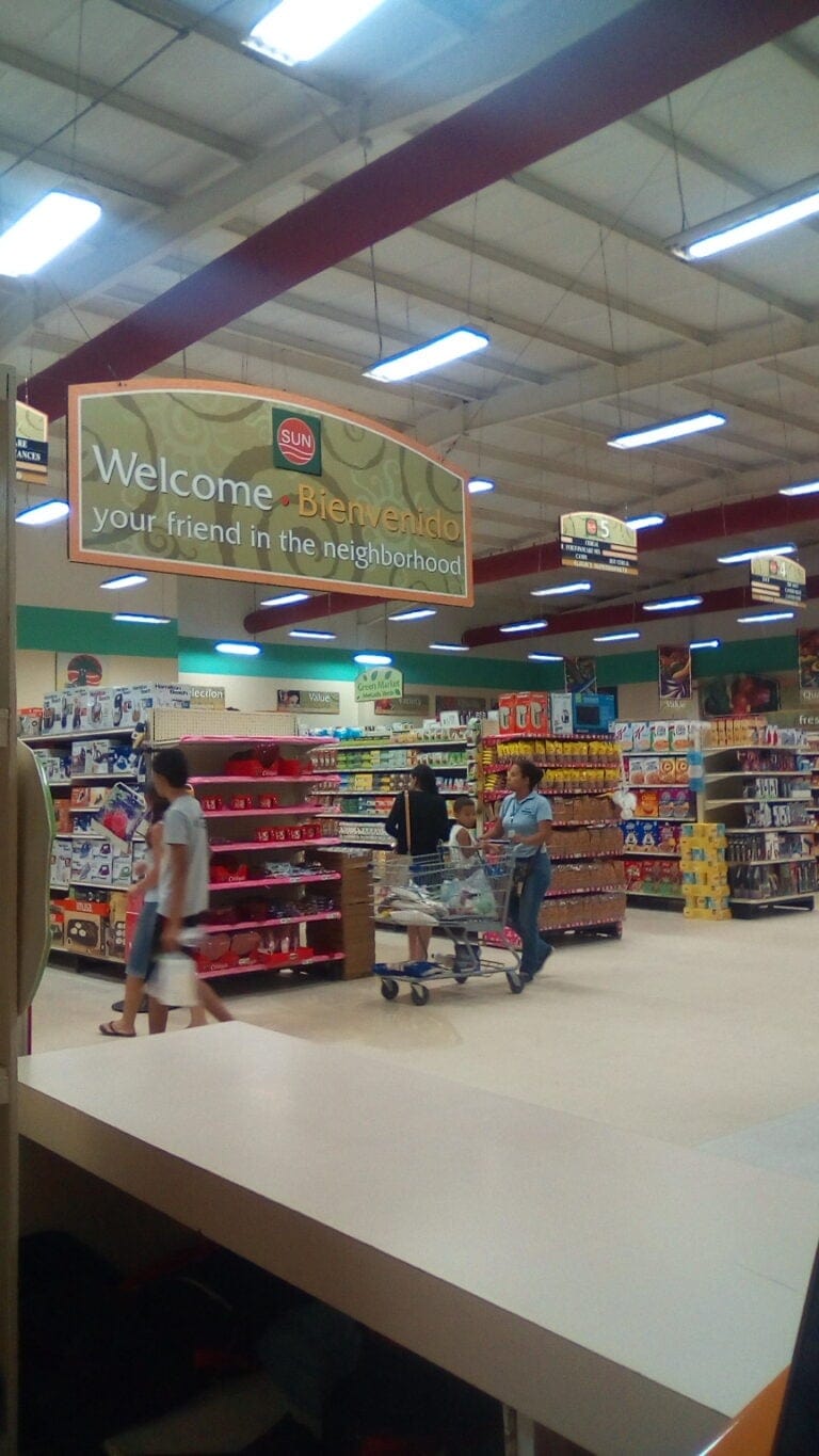 A family shops inside a large supermarket in Roatan, Honduras, with shelves stocked with groceries and household items and a sign welcoming customers in both English and Spanish.