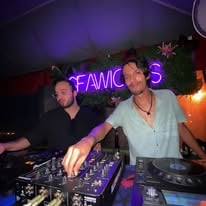 Two men are DJing at a nightclub in Roatan, Honduras, with a neon "FAMOUS" sign and colorful lighting creating a lively atmosphere.