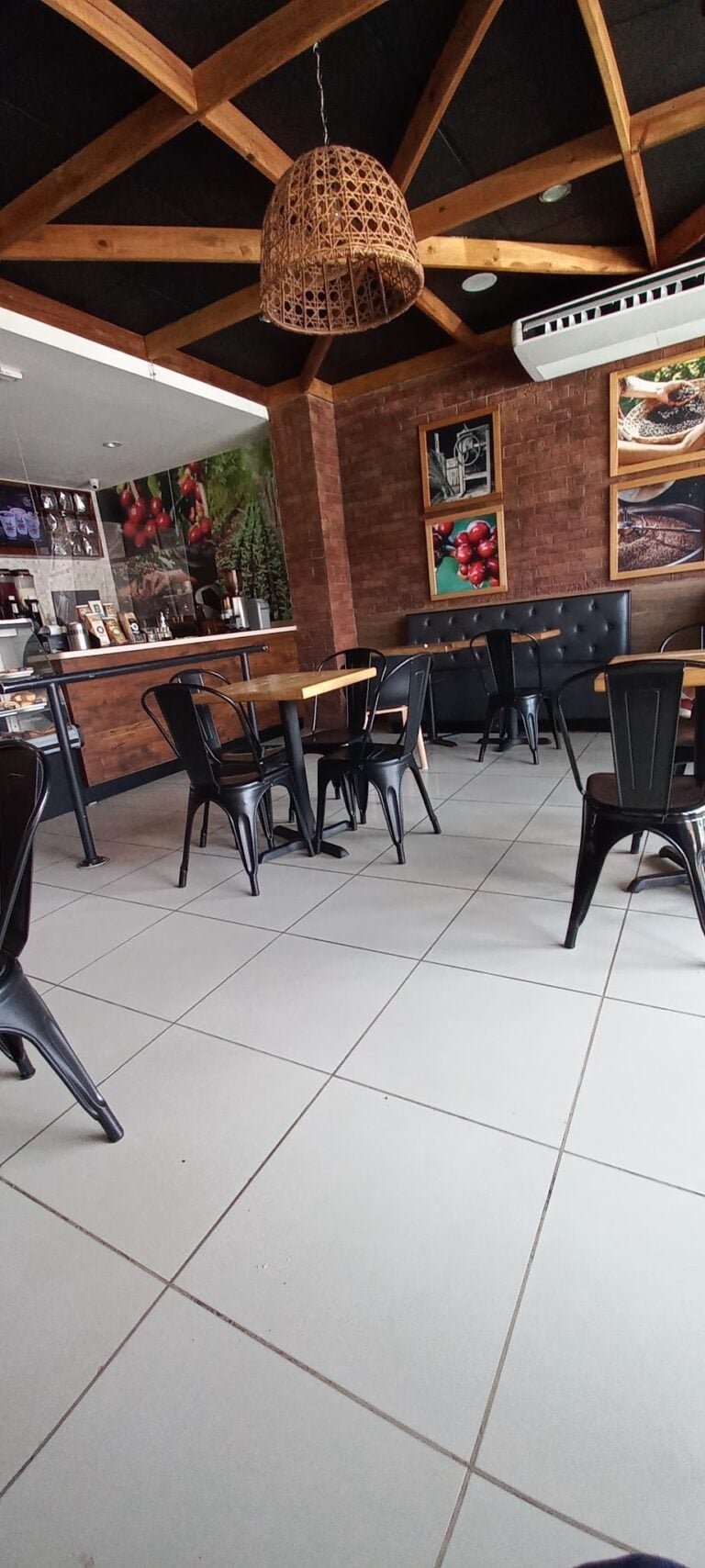 The interior of a café in Roatan, Honduras, features wooden beam ceilings, black metal chairs around wooden tables, a brick wall with framed pictures of coffee and harvest scenes, and a counter area displaying coffee products and equipment.