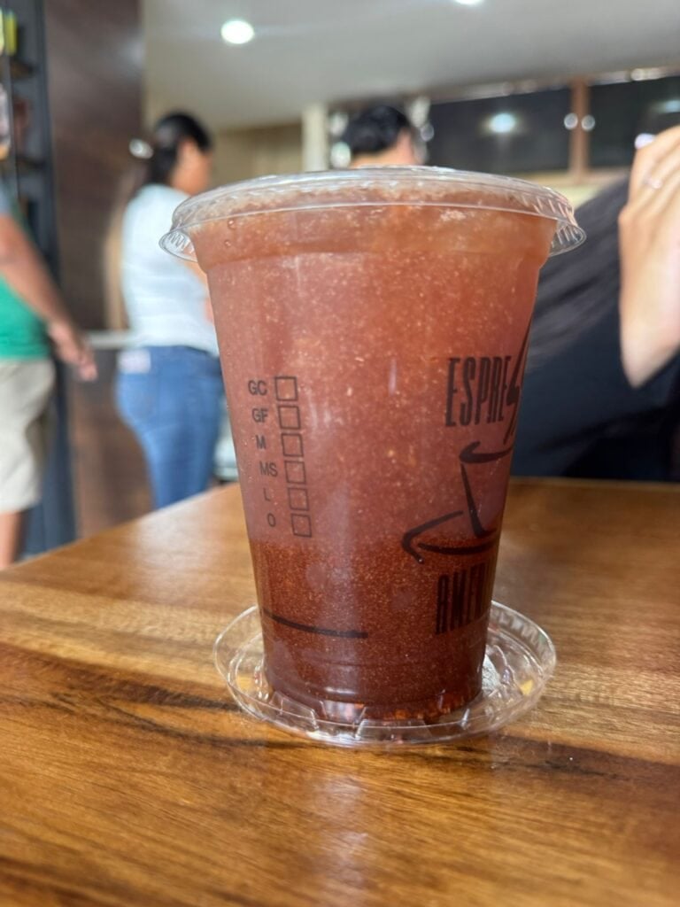A large iced blended beverage with a reddish-brown color sits on a wooden table inside a cafe or restaurant in Roatan, Honduras, with blurred customers and staff visible in the background.