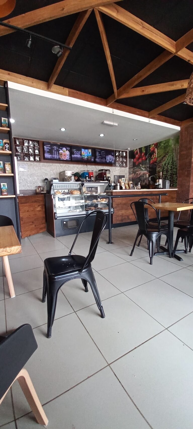 The image shows the interior of a cafe or coffee shop in Roatan, Honduras, featuring a wooden ceiling with exposed beams, a counter with a display case of baked goods, a menu board overhead, and seating with black metal chairs and wooden tables.