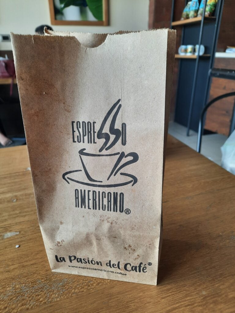 A close-up of a brown paper coffee bag with the Espresso Americano logo and the phrase "La Pasión del Café" on a wooden table inside a coffee shop in Roatan, Honduras.