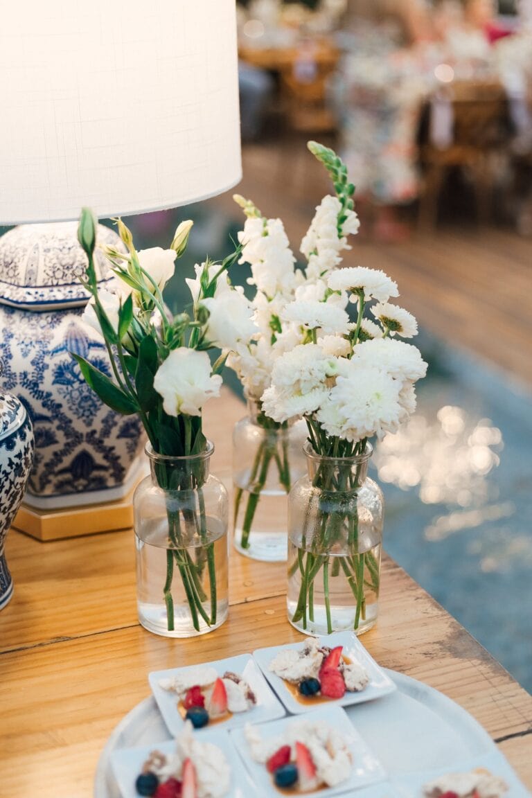 Vases with white flowers are displayed on a wooden table beside a lit lamp, with a background of a blurred dining or market area in Roatan, Honduras.