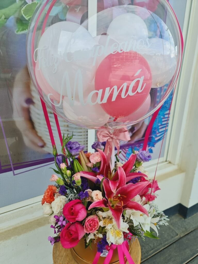 A colorful bouquet of pink lilies, roses, and purple and white flowers, with a pink ribbon, is topped with a transparent balloon that has "Feliz Cumpleaños Mamá" written on it, displayed outside a business on Roatan, Honduras.