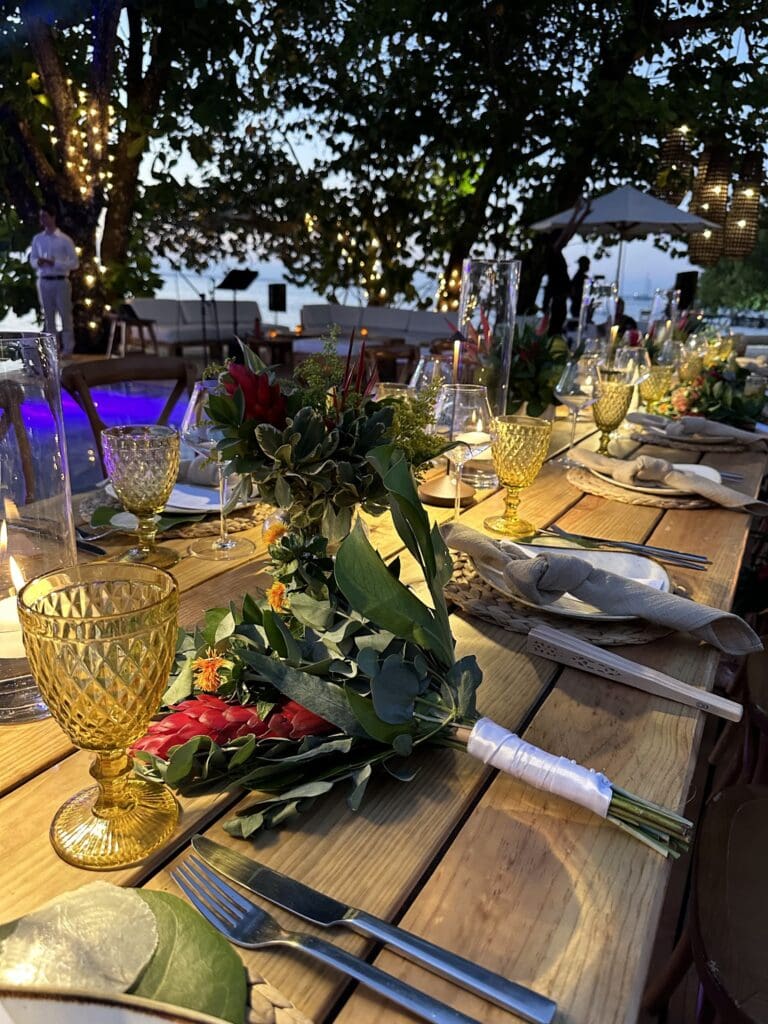 A decorated outdoor dining table with floral arrangements, glassware, and cutlery set under string lights and a large tree, with a view of the ocean in Roatan, Honduras.