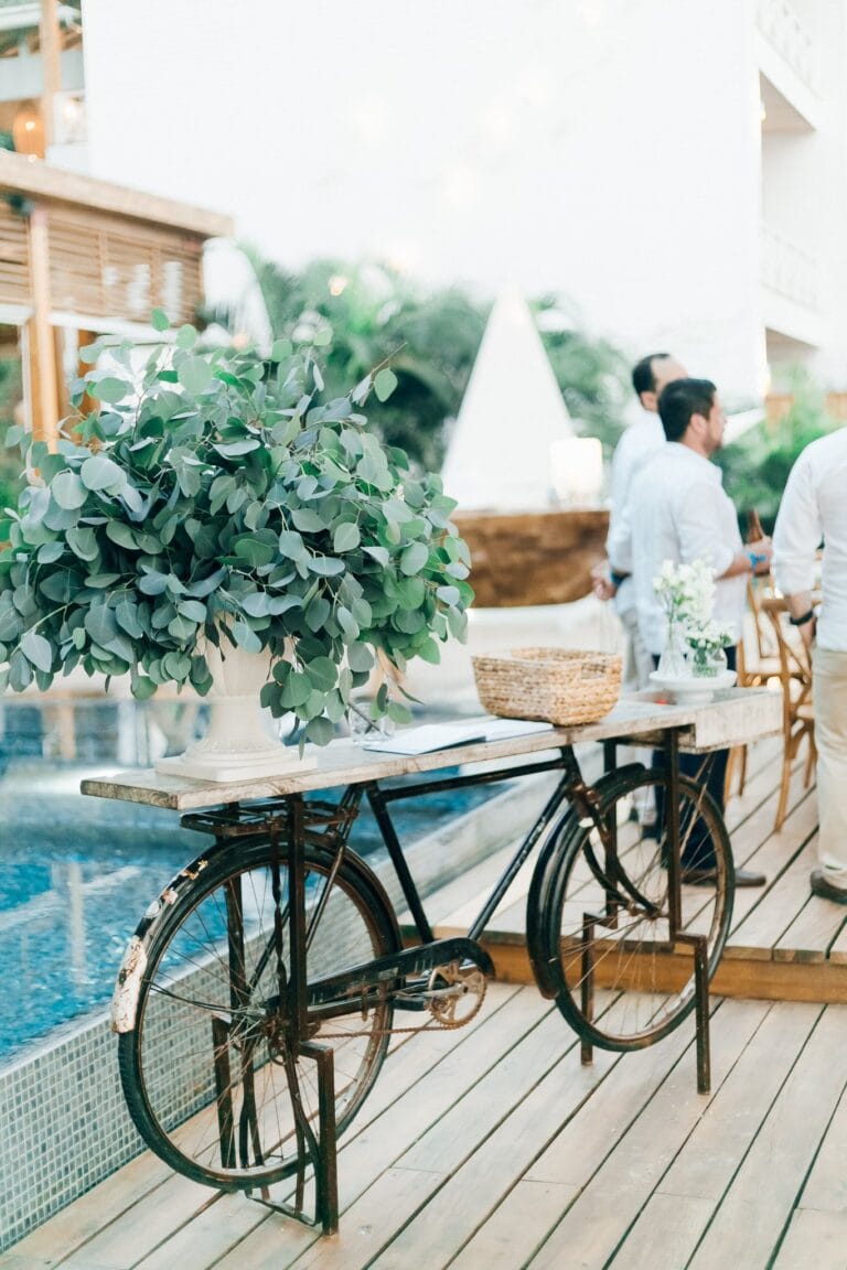 A vintage bicycle with a large white planter filled with green foliage is displayed on a wooden deck at a restaurant or cafe in Roatan, Honduras, with people dining in the background and tropical greenery visible.