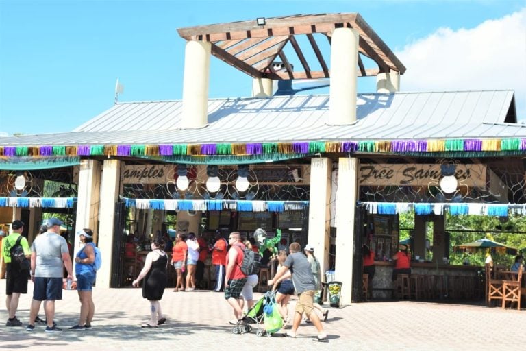 A lively crowd gathers outside a colorful bar in Roatan, Honduras, featuring a decorated awning with purple, green, and gold fringe, and a sign indicating free sangria and samples, with the building's open interior and people enjoying drinks and socializing on a sunny day.