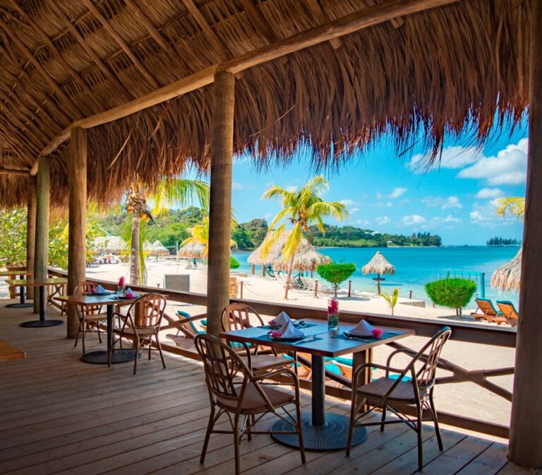 Beachfront restaurant on Roatan with a thatched roof, wooden tables set with napkins and flowers, and a view of sandy beach, palm trees, and turquoise Caribbean Sea in Honduras.