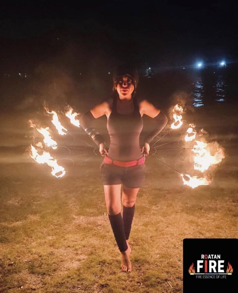 A woman performs a fire dance at night on a grassy area near the water on Roatan, Honduras, with flames connected by wires creating an illuminated circular pattern behind her.