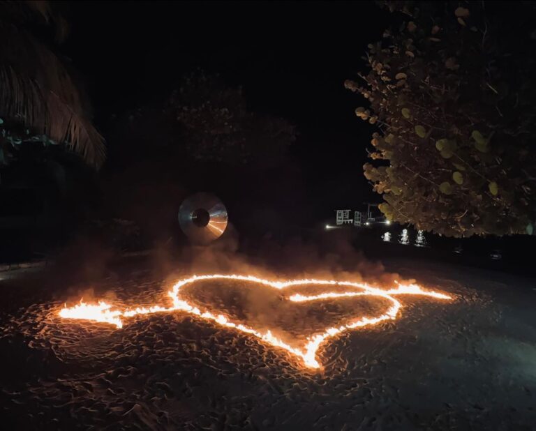 A flaming heart is drawn on the sand at night on a beach in Roatan, Honduras, with palm trees and a metal sculpture visible in the background.