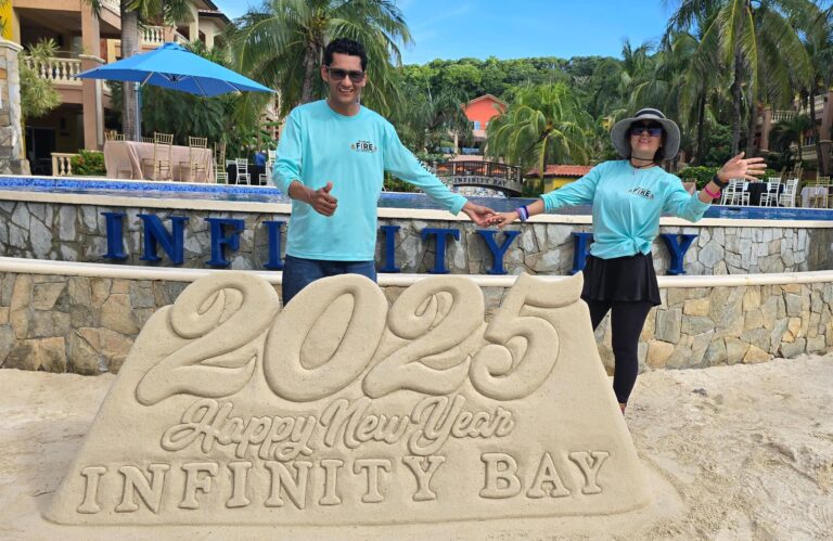 Two people in matching light blue shirts with "Roatan Fire" logos pose on a sandy beach in Roatan, Honduras, holding hands with a large sand sculpture of "Happy New Year" and "Infinity Bay" in front of them, surrounded by palm trees, resort buildings, and a pool area.
