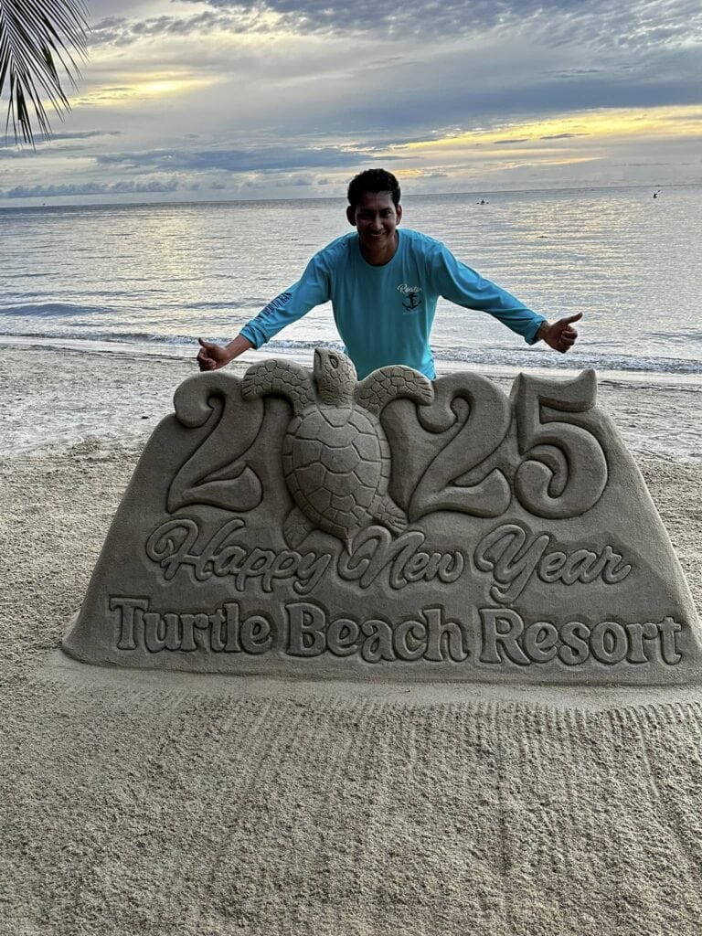 A man in a blue shirt stands behind a large sand sculpture at Turtle Beach Resort in Roatan, Honduras, with the ocean, boats, and a colorful sunset sky in the background.