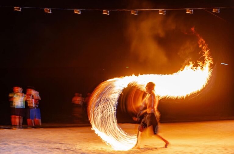 A fire performer creates a large arc of fire on a beach in Roatan, Honduras, during a nighttime event with a small group of spectators watching in the background.