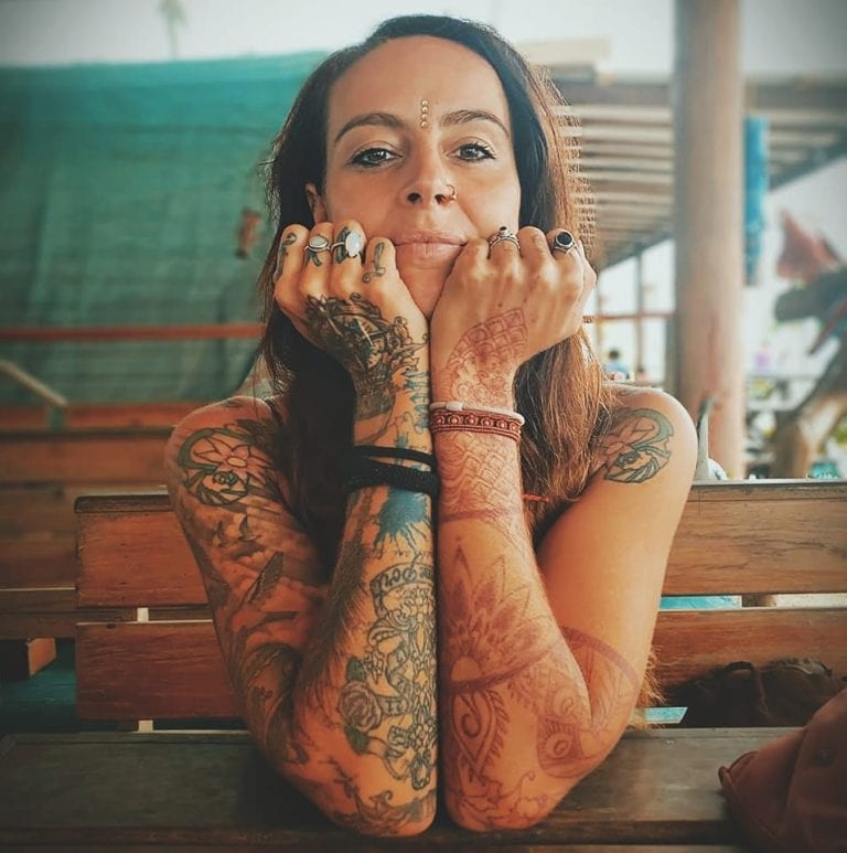 A tattooed woman with long hair, multiple rings, and henna designs on her hands and arms rests her chin on her hands while sitting at a wooden table in an outdoor setting on Roatan, Honduras, with a shaded area and colorful decor in the background.