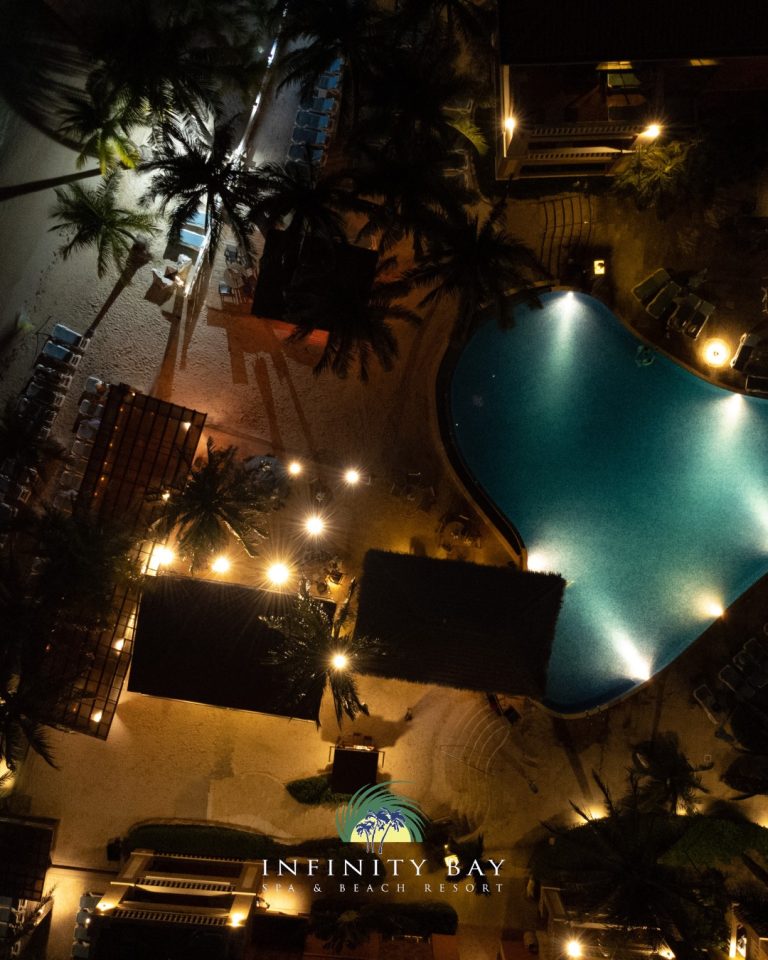 A nighttime aerial view of Infinity Bay Spa & Beach Resort on Roatan, Honduras, showcasing a large illuminated swimming pool surrounded by palm trees, lounge chairs, and shaded cabanas.