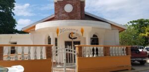 A white church with a brick steeple, decorative columns, and a "Welcome" sign featuring a cross, flames, and a Bible, located in Roatan, Honduras.